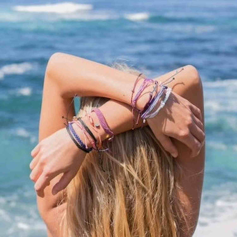 Back of woman's head with water in background. Hands over head with Pura Vida bracelets on wrist.