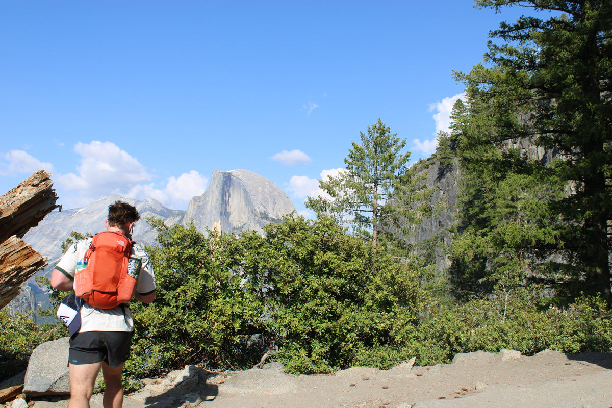 Person with a red backpack hiking in a mountainous area with trees and clear sky.